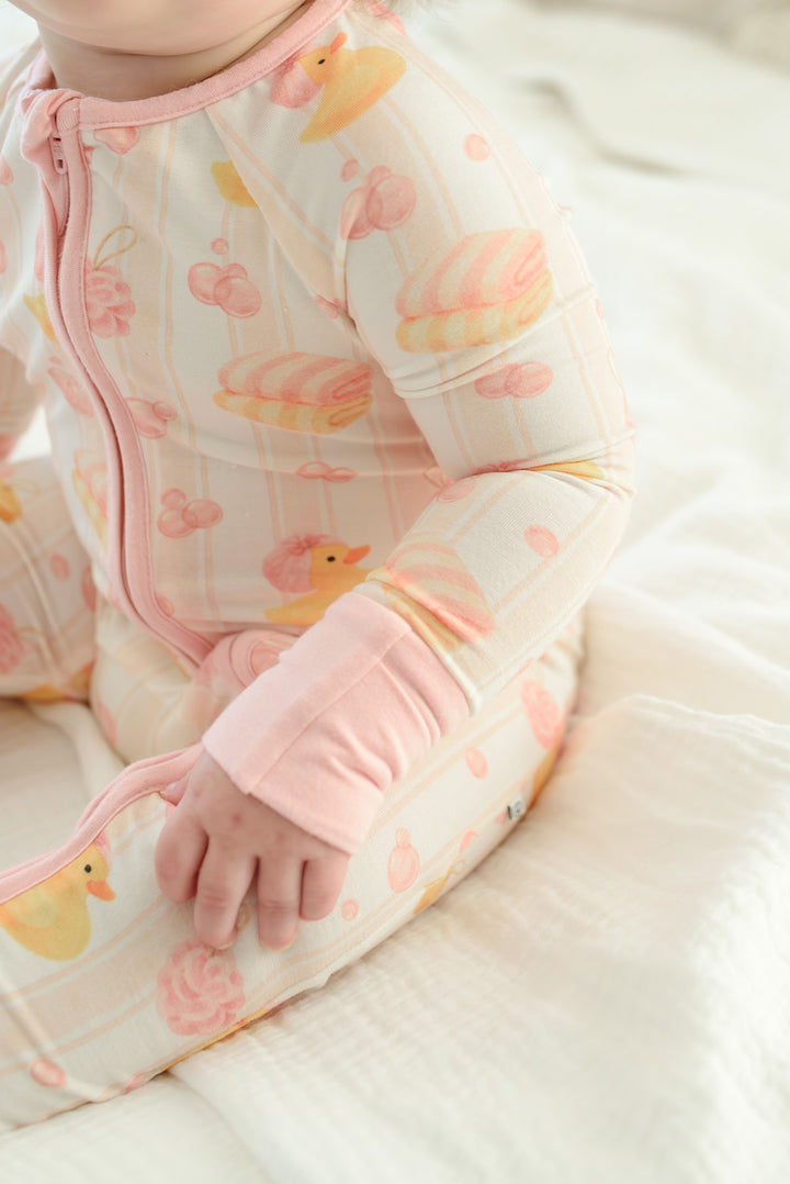 Happy baby girl wearing pink Bubble Duckies zip romper sitting on rubber duck print bedding with pillow background