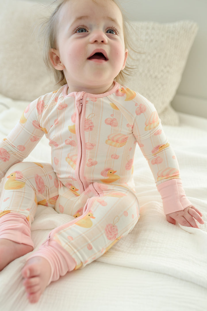 Baby girl wearing pink Bubble Duckies zip romper on rubber duck print bedding with pink bow hair accessory