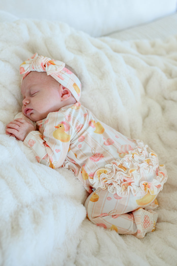 Sleeping newborn in pink Bubble Duckies ruffle footie pajamas with bow headwrap on white fur blanket
