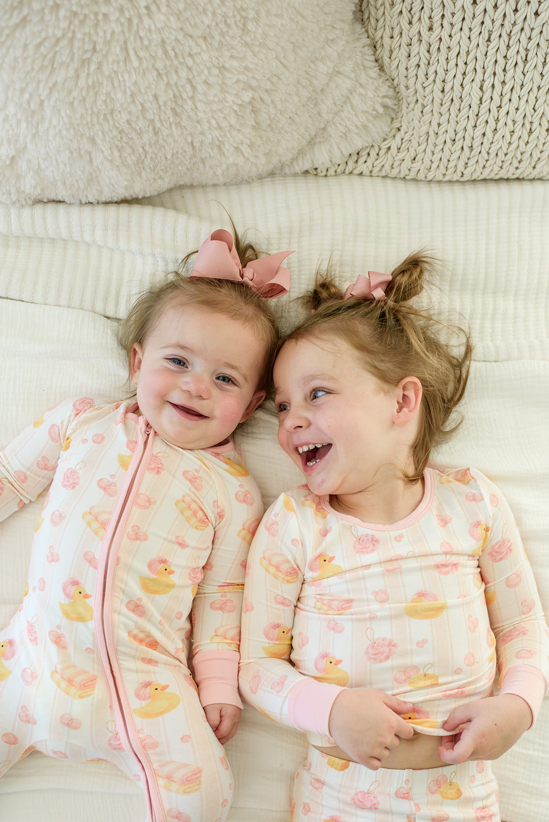 Smiling baby girl in pink Bubble Duckies zip romper pajamas with bow on matching bubble duck print bedding