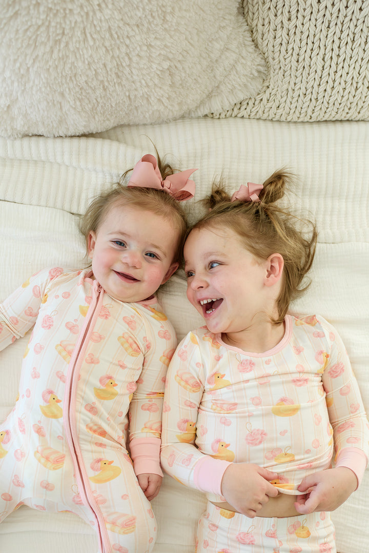 Smiling baby girl in pink Bubble Duckies zip romper pajamas with bow on matching bubble duck print bedding