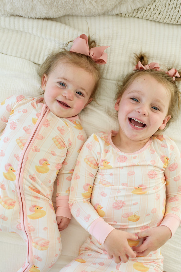 Two sisters lying on bed wearing matching pink Bubble Duckies pajamas - baby in zip romper and older girl in two-piece set