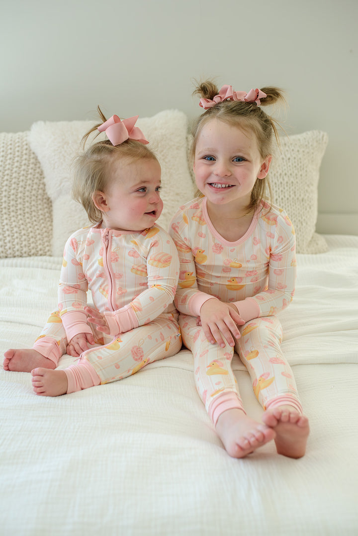 Two sisters sitting together on bed wearing matching pink Bubble Duckies bamboo pajamas with yellow rubber duck print