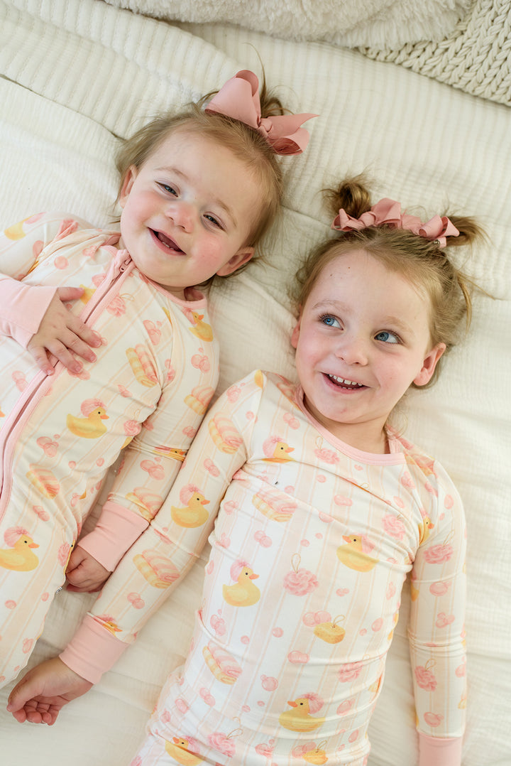 Two sisters lying on bed in matching pink Bubble Duckies bamboo pajamas with yellow rubber duck print