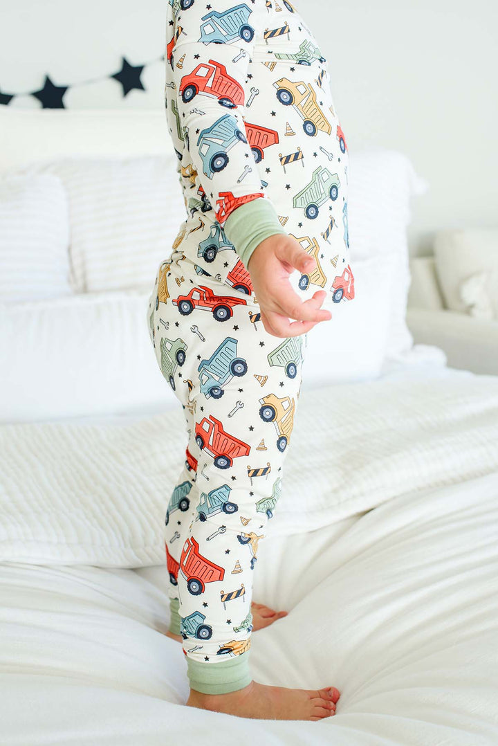Side view of a toddler standing barefoot on a white bed wearing Caden Lane cream knit pajamas in a Busy Builder print featuring dump trucks and barricades.