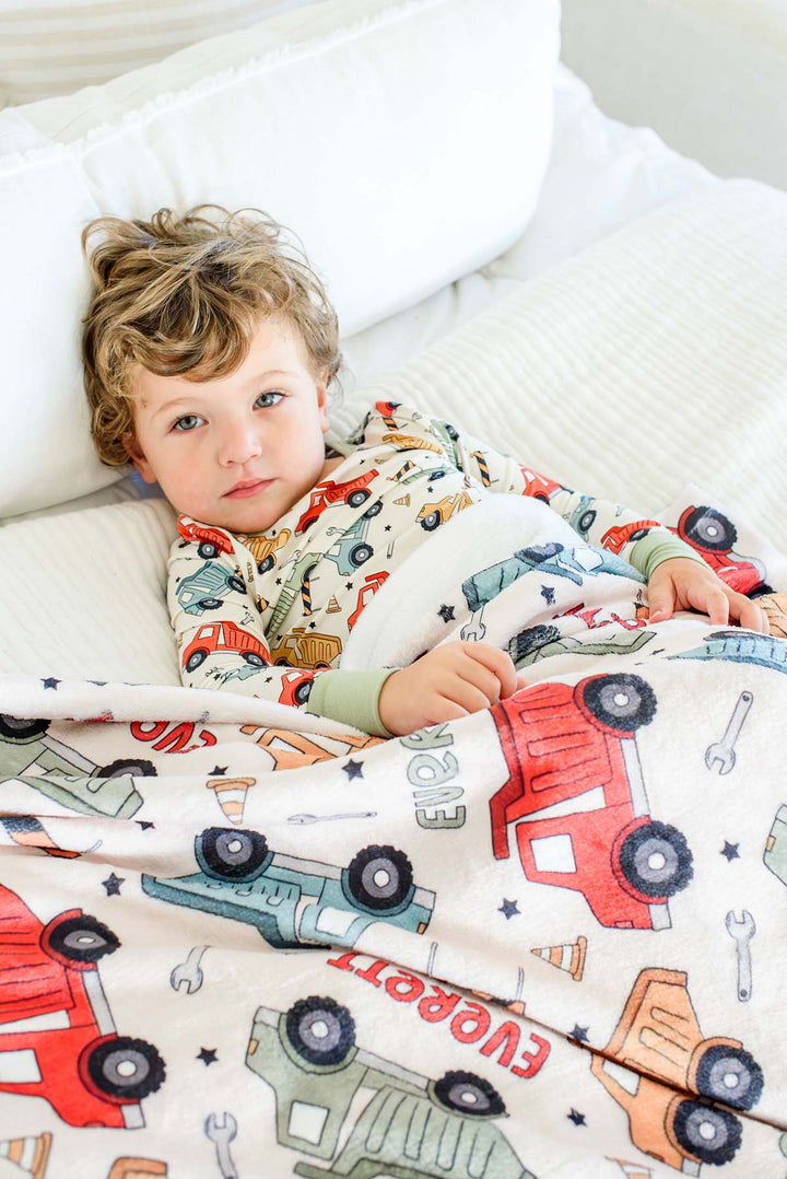 A curly-haired toddler propped against white pillows under a Caden Lane cream plush blanket in a Busy Builder print featuring dump trucks and cones, reading "Everett," paired with matching pajamas.