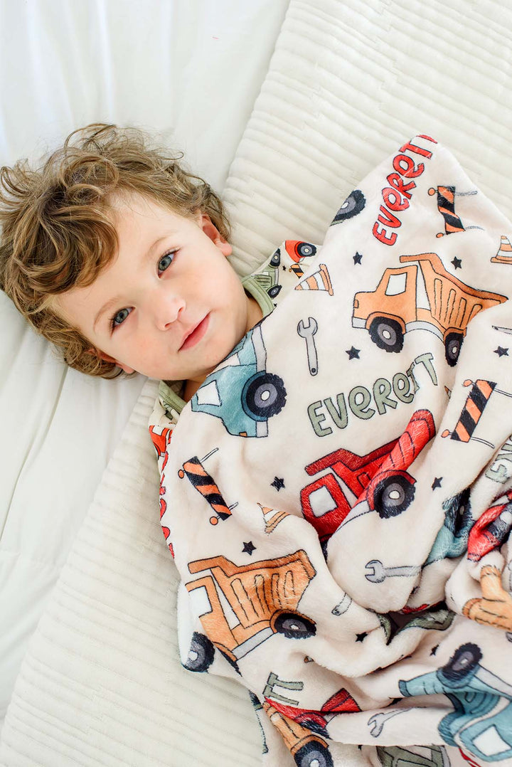 Overhead shot of a curly-haired toddler tucked to the chin under a Caden Lane cream plush blanket in a Busy Builder print featuring dump trucks and wrenches, reading "Everett."