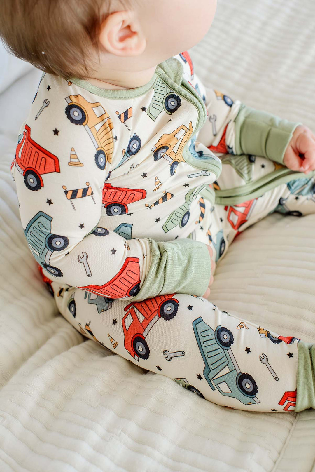 Over-the-shoulder view of a baby sitting on a white quilt, wearing a Caden Lane cream knit zip romper in a Busy Builder print featuring dump trucks, with sage cuffs.