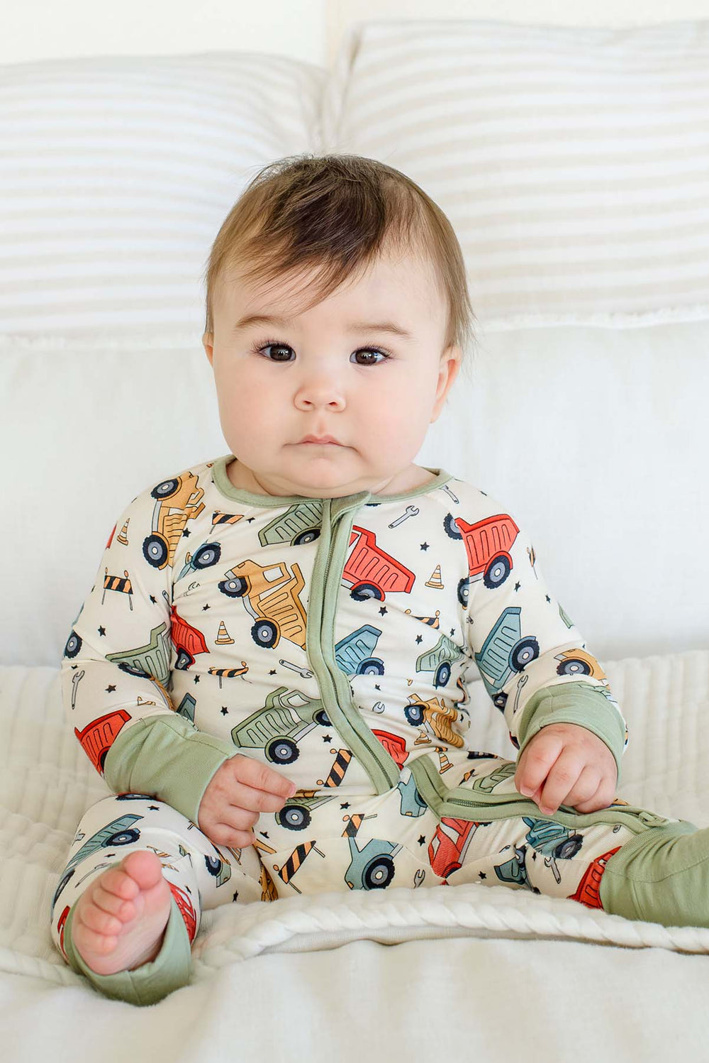 A brown-eyed baby sitting up against white pillows, wearing a Caden Lane cream knit zip romper in a Busy Builder print featuring dump trucks and cones, with sage trim.