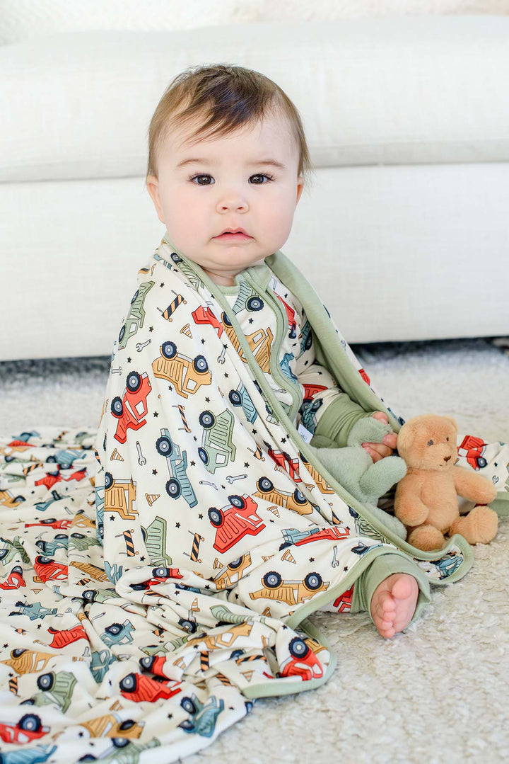 A baby on a shag rug with a Caden Lane cream knit blanket around the shoulders in a Busy Builder print featuring dump trucks, paired with a teddy bear.