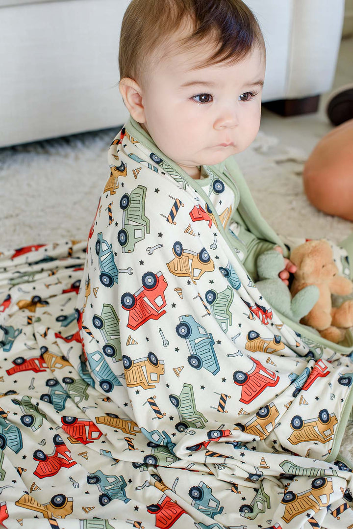 A baby gazing sideways, wrapped in a Caden Lane cream knit blanket with sage trim in a Busy Builder print featuring dump trucks, paired with plush toys.