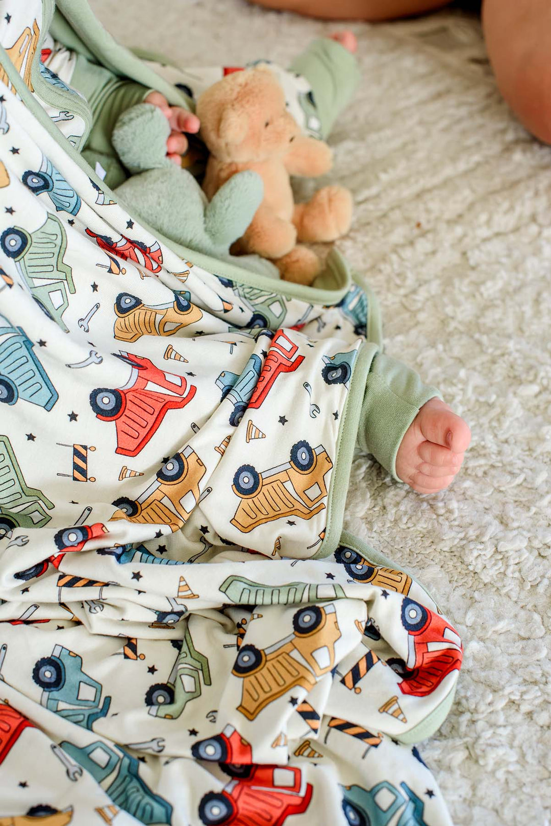 Close-up of a baby's fist and a teddy bear peeking from a Caden Lane cream knit blanket with sage trim in a Busy Builder print featuring dump trucks and barricades.