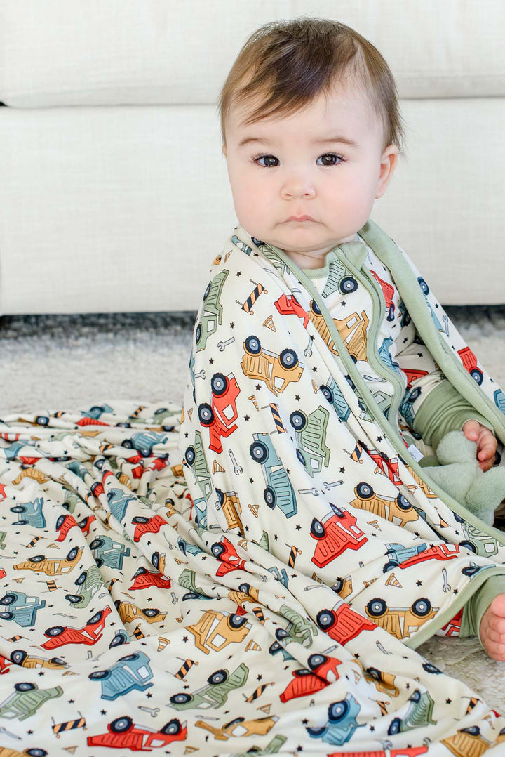 A baby looking at the camera with a Caden Lane cream knit blanket pooled around them in a Busy Builder print featuring dump trucks, paired with a matching sleeper.