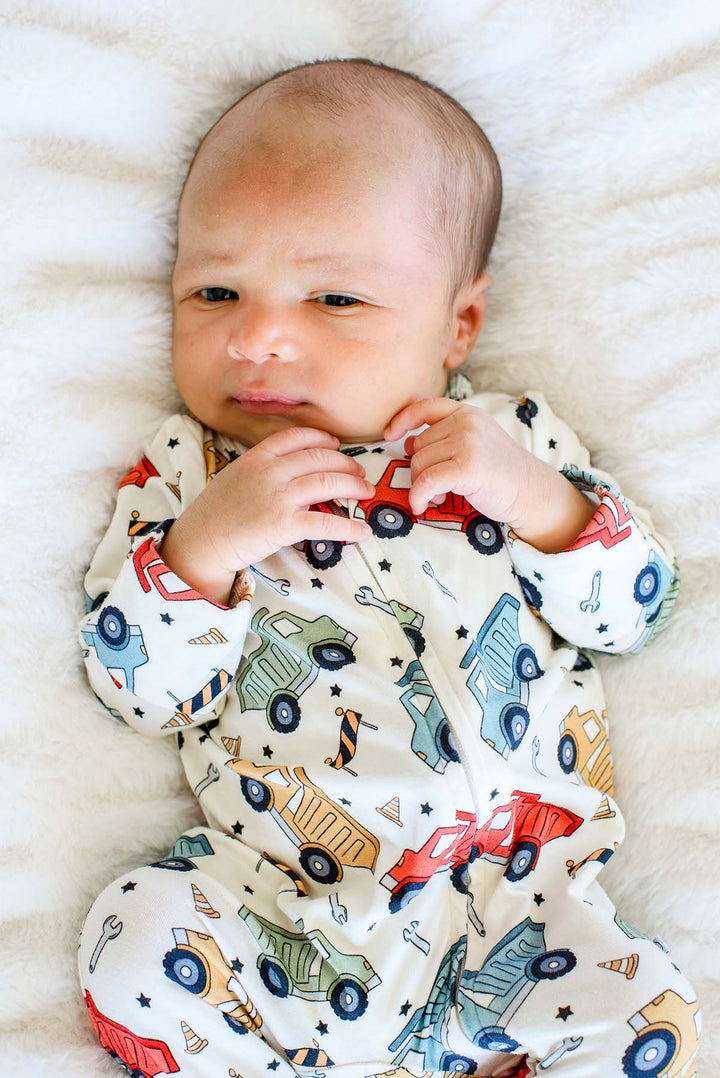 A newborn lying on white faux fur with hands tucked near the chin, wearing a Caden Lane cream knit footed sleeper in a Busy Builder print featuring dump trucks.
