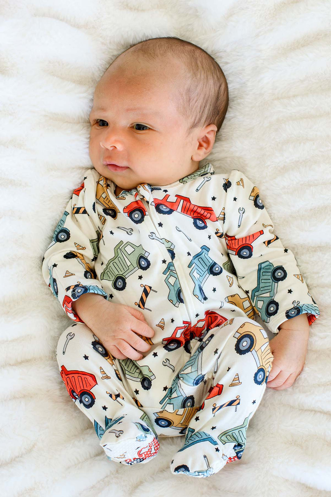 Full-length shot of a newborn glancing sideways with knees bent, wearing a Caden Lane cream knit footed sleeper in a Busy Builder print featuring dump trucks and wrenches.