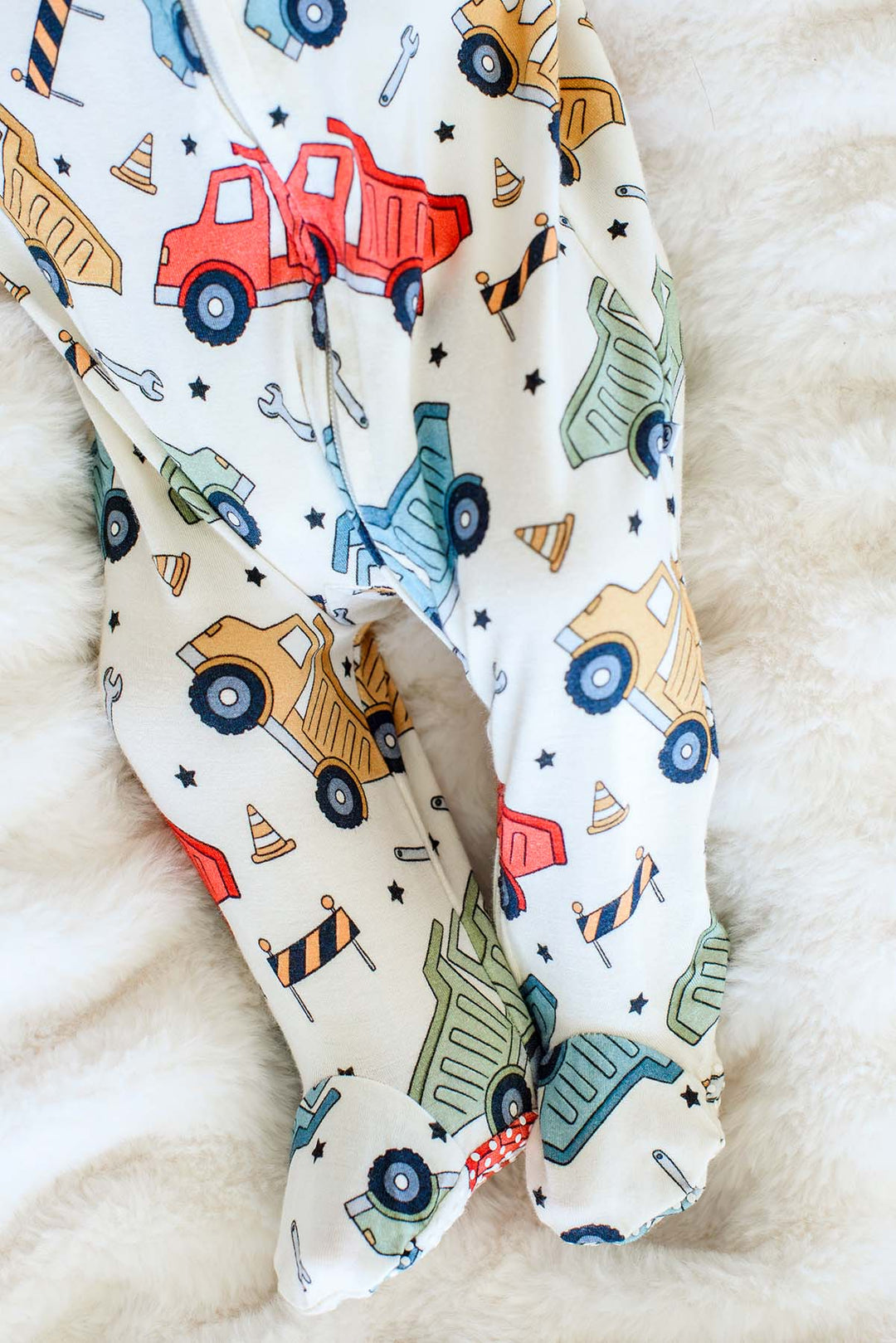 Close-up of the footed legs of a Caden Lane cream knit sleeper in a Busy Builder print featuring dump trucks and cones, with red polka-dot grippy soles.