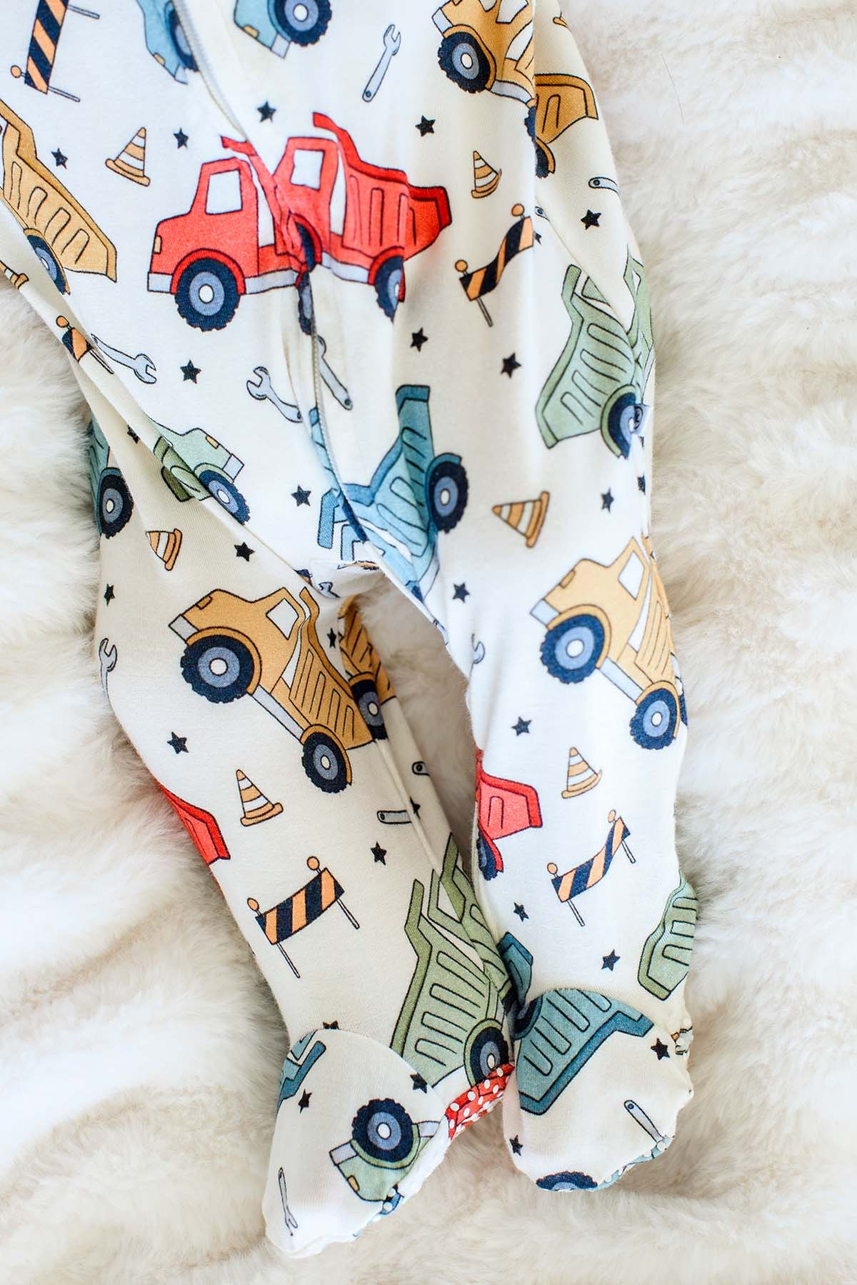Close-up of the footed legs of a Caden Lane cream knit sleeper in a Busy Builder print featuring dump trucks and cones, with red polka-dot grippy soles.