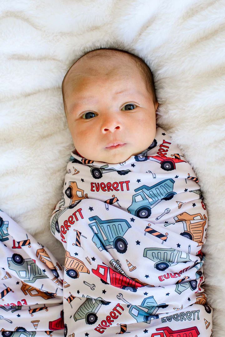 Close-up of a wide-eyed newborn looking at the camera, wrapped in a Caden Lane cream swaddle in a Busy Builder print featuring dump trucks and barricades, reading "Everett."
