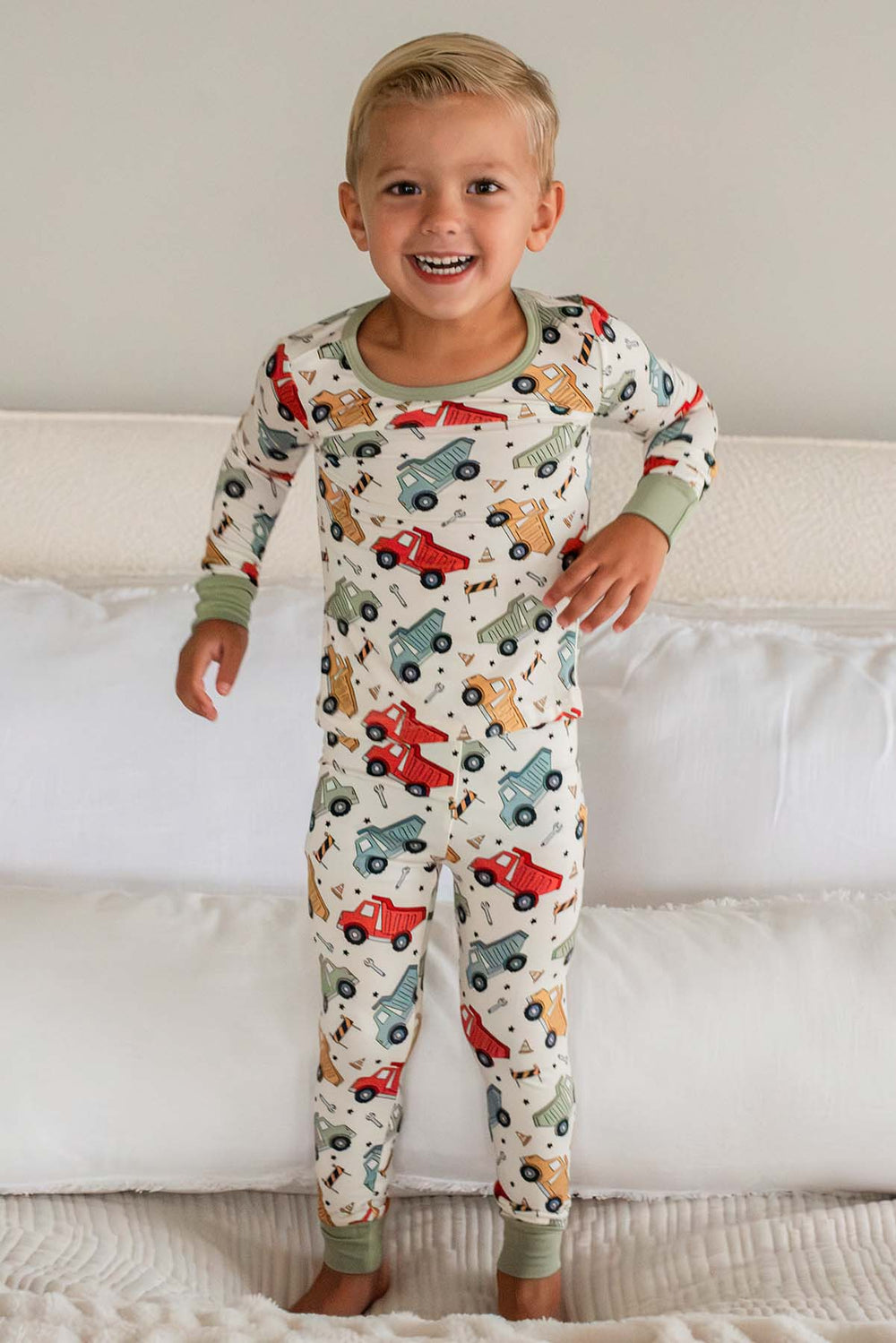 A grinning blond toddler standing on a bed wearing a Caden Lane cream knit pajama set in a Busy Builder print featuring dump trucks, with sage cuf