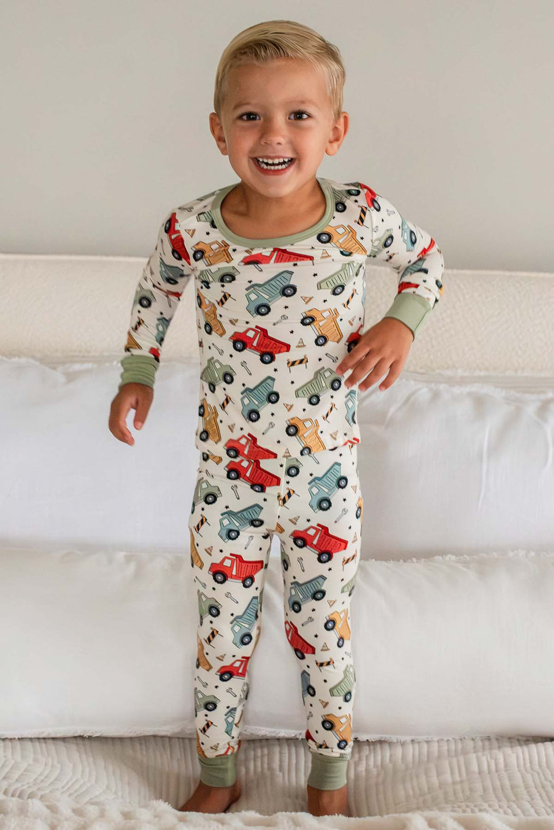 A grinning blond toddler standing on a bed wearing a Caden Lane cream knit pajama set in a Busy Builder print featuring dump trucks, with sage cuf