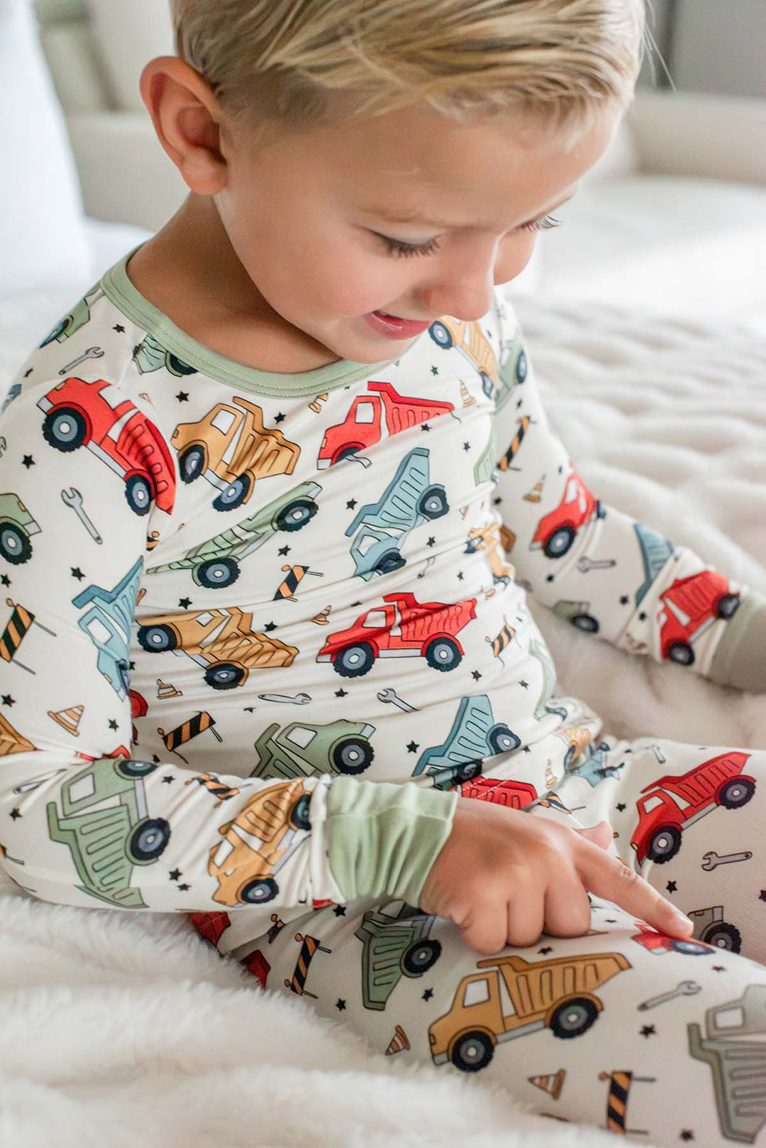A blond toddler pointing at a truck on his pant leg, wearing a Caden Lane cream knit pajama set in a Busy Builder print featuring dump trucks and barricades.