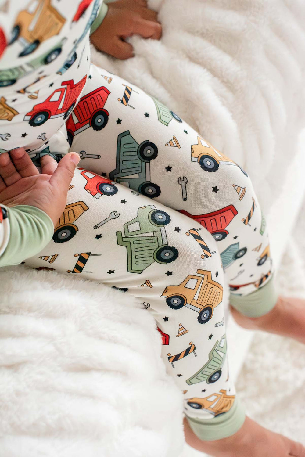 Close-up of a toddler's bent legs in Caden Lane cream knit pajama pants in a Busy Builder print featuring dump trucks and cones, with sage ankle cuffs.