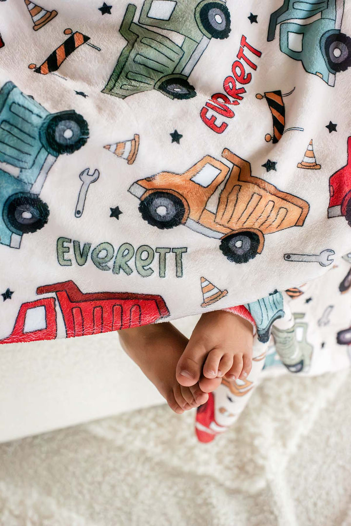Close-up of a toddler's bare feet peeking out beneath a Caden Lane cream plush blanket in a Busy Builder print featuring dump trucks, cones, and barricades, reading "Everett."