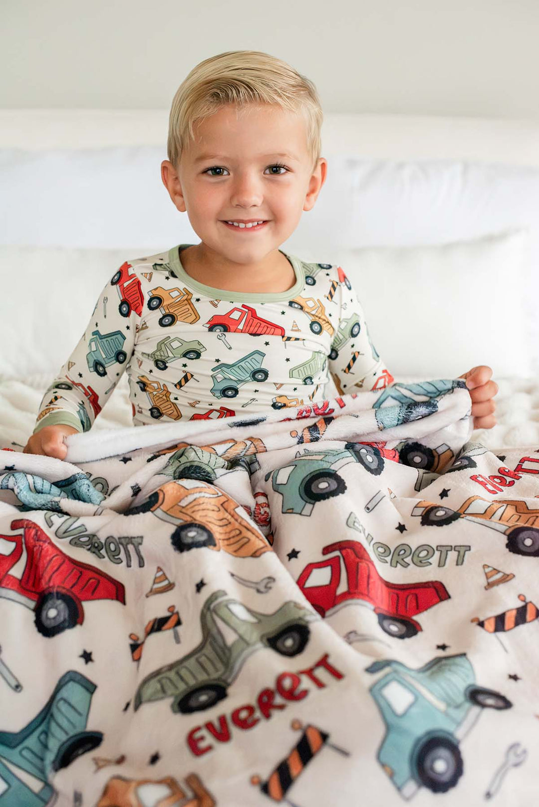 A smiling blond boy sitting with a Caden Lane cream plush blanket over his lap in a Busy Builder print featuring dump trucks, reading "Everett," paired with matching pajamas.