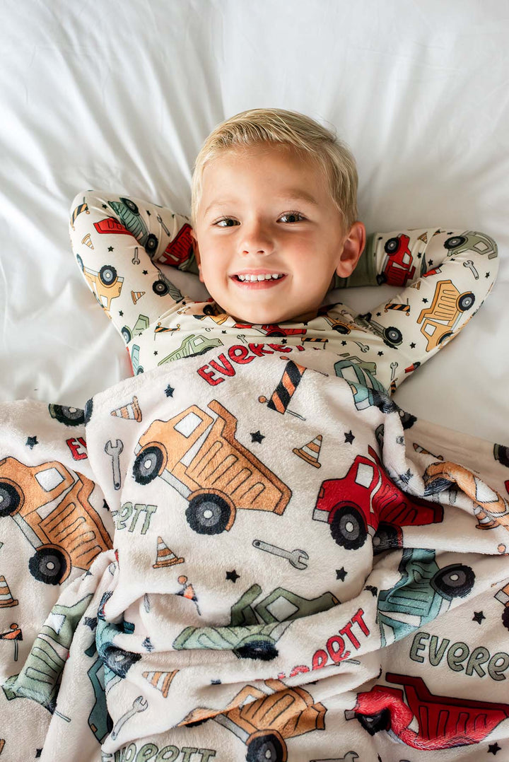 A blond boy lying with hands behind his head under a Caden Lane cream plush blanket in a Busy Builder print featuring dump trucks, reading "Everett," paired with matching pajamas.