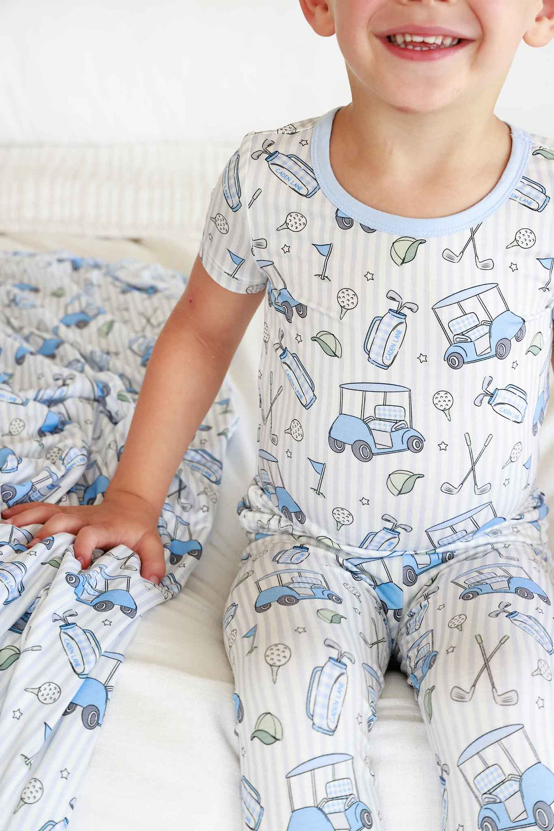 boy sitting on bed in caddie crew blue two piece pajama set with matching blanket next to him 