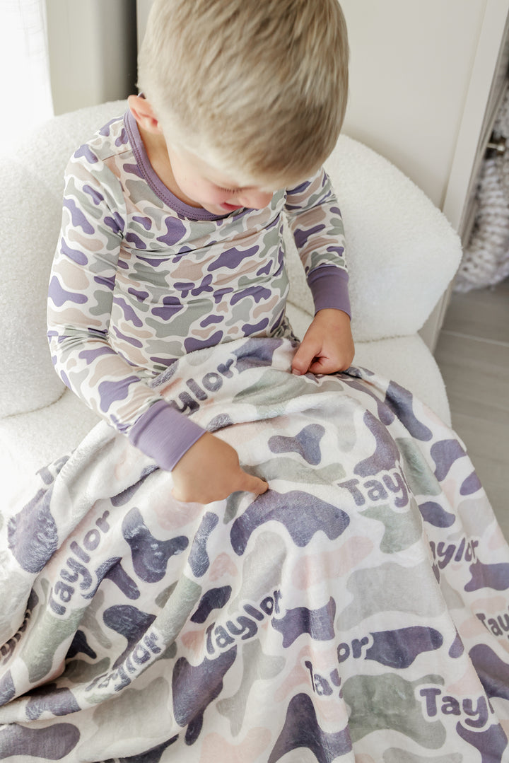 Toddler boy in camo print pajamas sitting and looking at his personalized camo cutie kids blanket with custom name