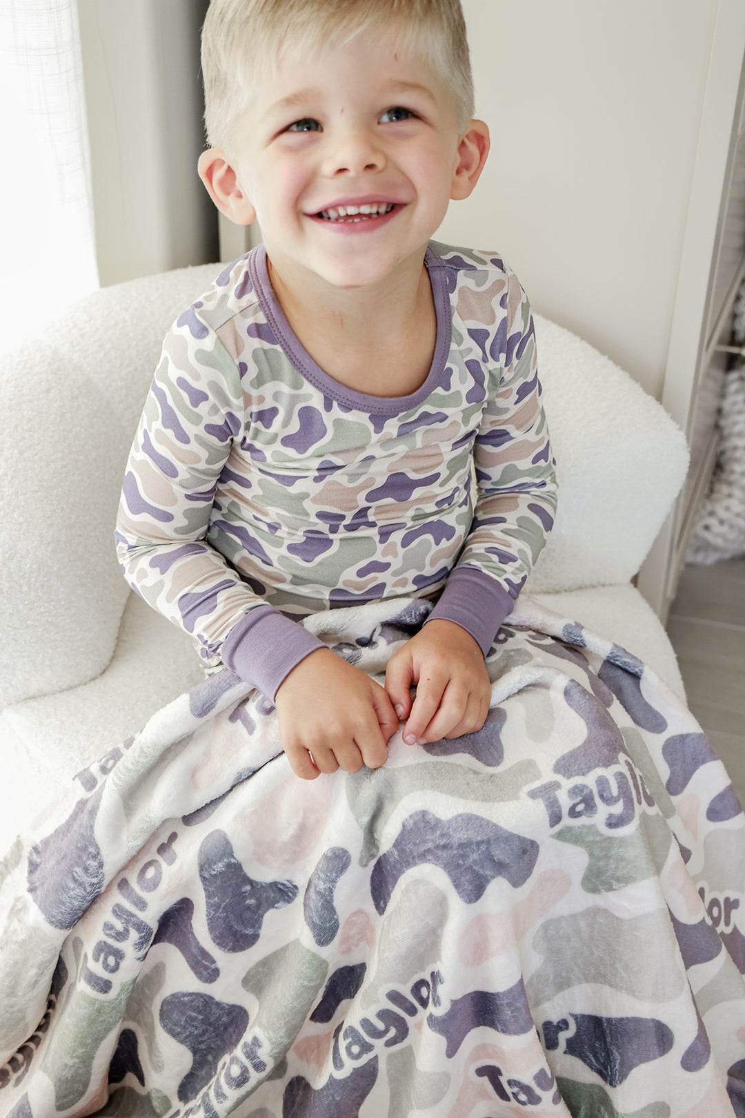 Smiling toddler boy in camo print pajamas holding a personalized camo cutie kids blanket with custom name