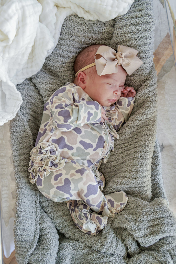 Newborn baby girl sleeping in a camo cutie print ruffle footie with a taupe ribbon bow headband on a gray knit blanket
