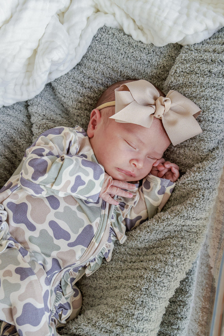 Sleeping newborn baby girl in a camo cutie print ruffle zipper footie with a taupe ribbon bow headband on a gray blanket