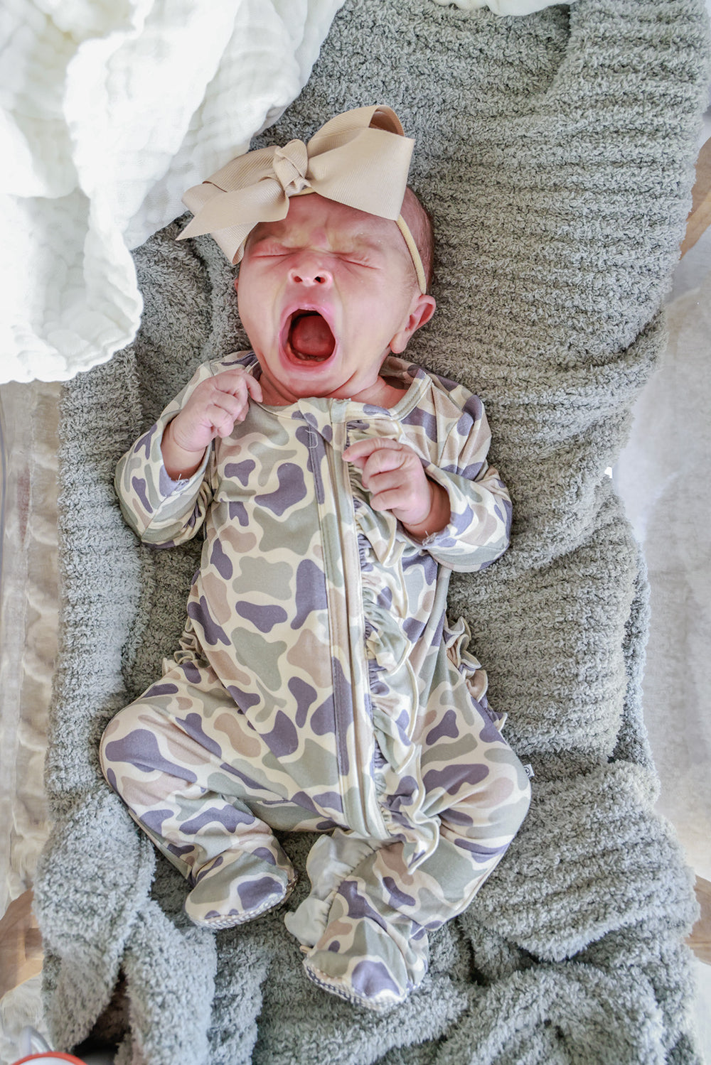 Newborn baby wearing a camo cutie print footie and taupe ribbon bow headband, yawning on a gray textured blanket