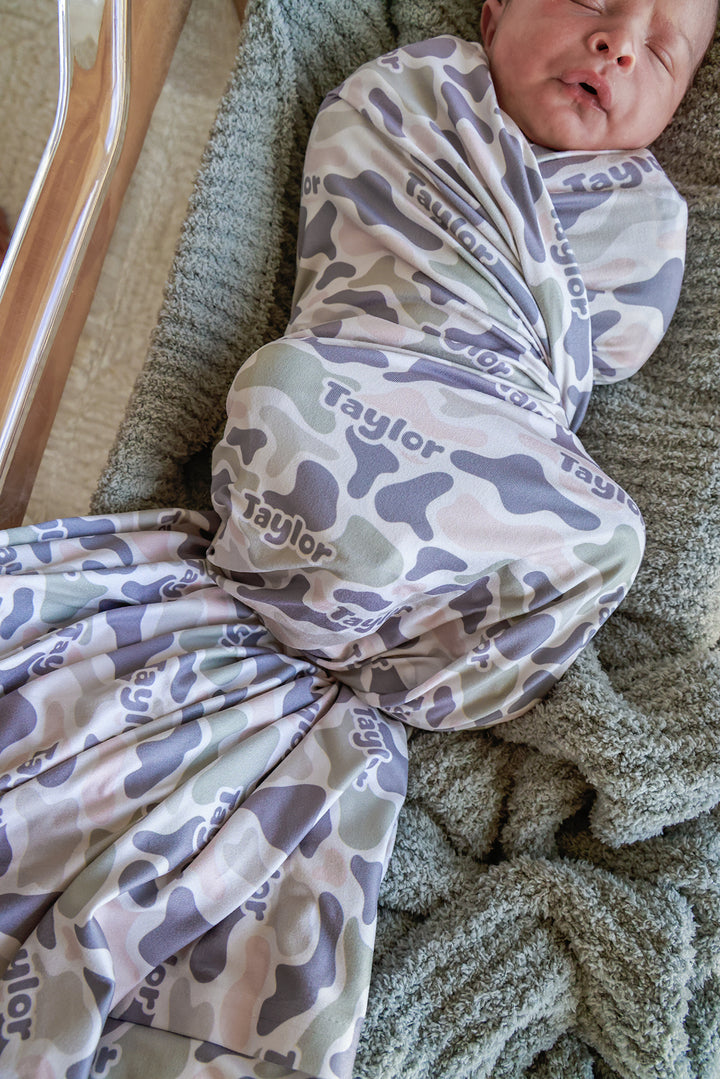 Newborn loosely swaddled in personalized camo cutie blanket in a bassinet, with arms showing and custom name visible