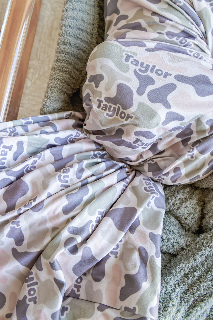 Close-up of personalized camo cutie swaddle blanket bunched and knotted on a gray blanket, showing custom name in camo print