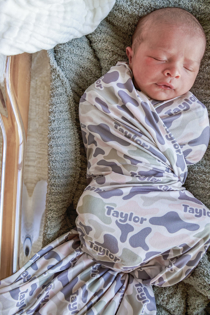 Sleeping newborn swaddled in a personalized camo cutie swaddle blanket with custom name prominently displayed