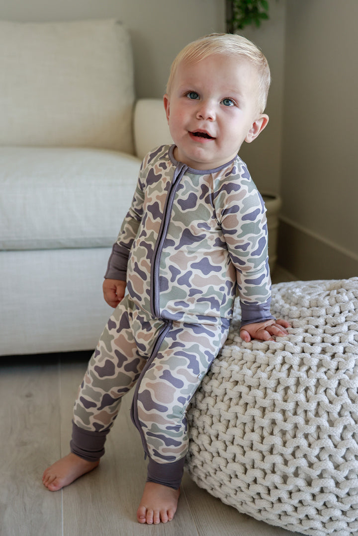 Toddler boy standing in a camo cutie convertible zip romper with camo print and gray cuffs