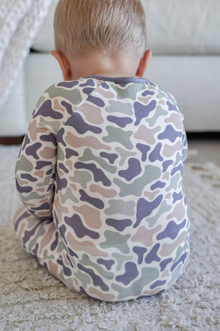 Toddler boy looking forward in camo cutie convertible zip romper, leaning on a textured pouf