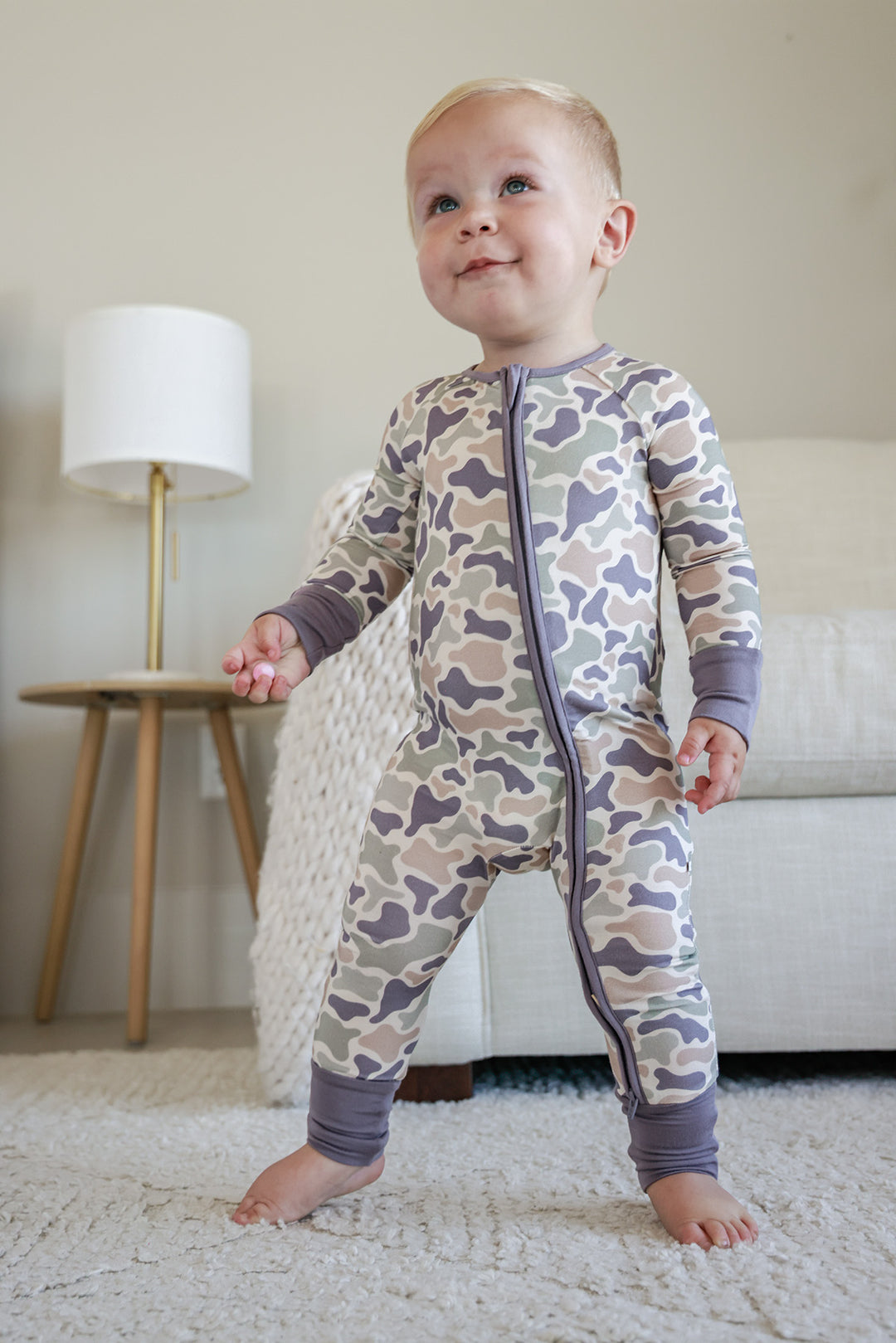 Baby in camo cutie convertible zip romper on rug from behind