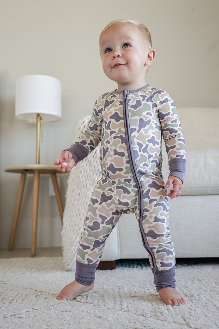 Baby in camo cutie convertible zip romper on rug from behind