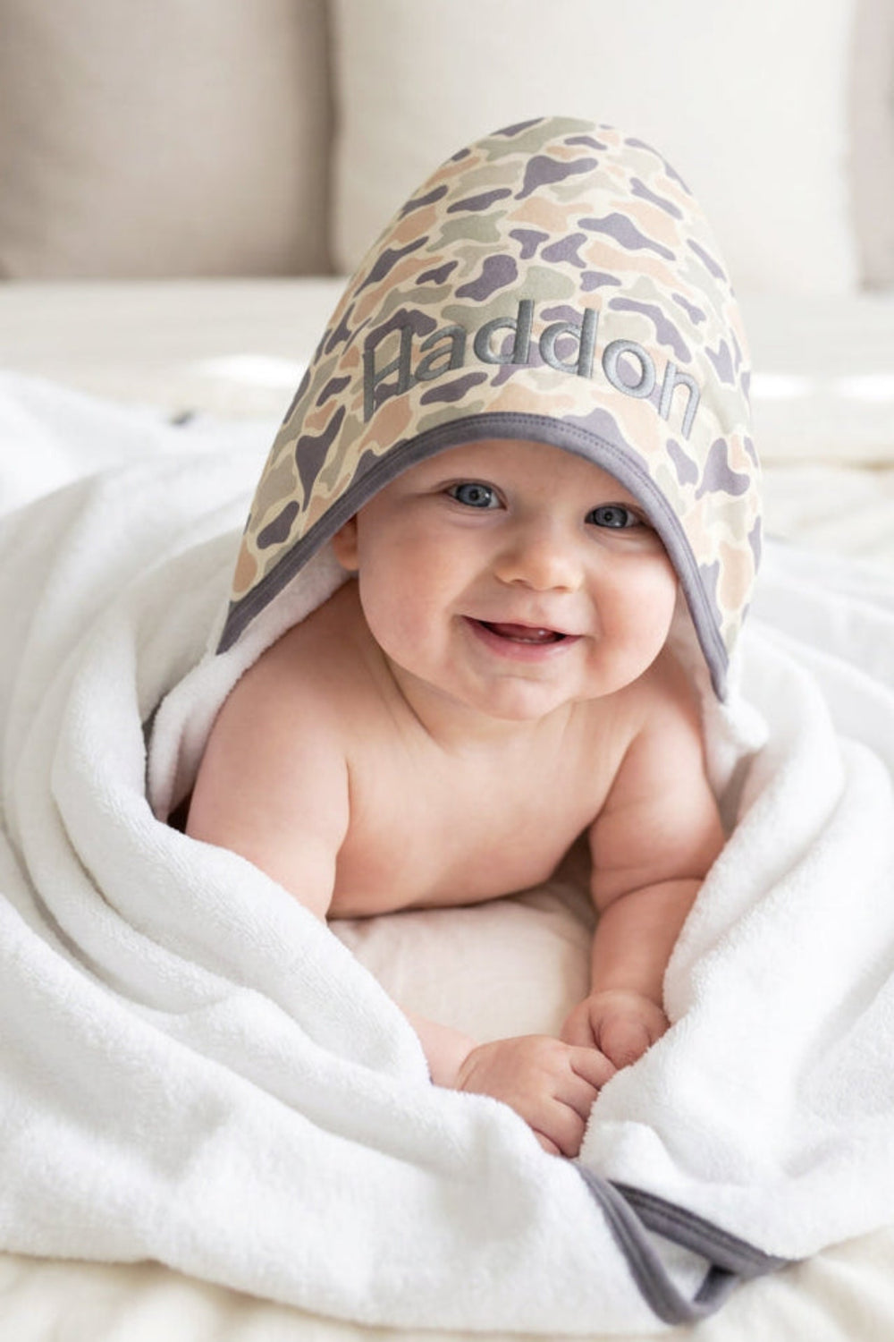 Baby lying down wrapped in an embroidered bamboo hooded towel with a camo print hood in tan and brown