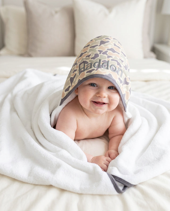 Baby lying down wrapped in an embroidered bamboo hooded towel with a camo print hood in tan and brown