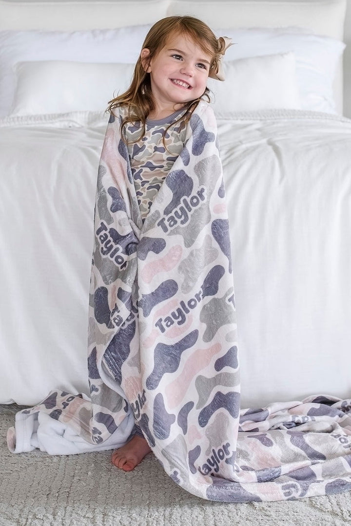 Smiling girl wrapped in a personalized camo cutie blanket with custom name, standing next to a bed