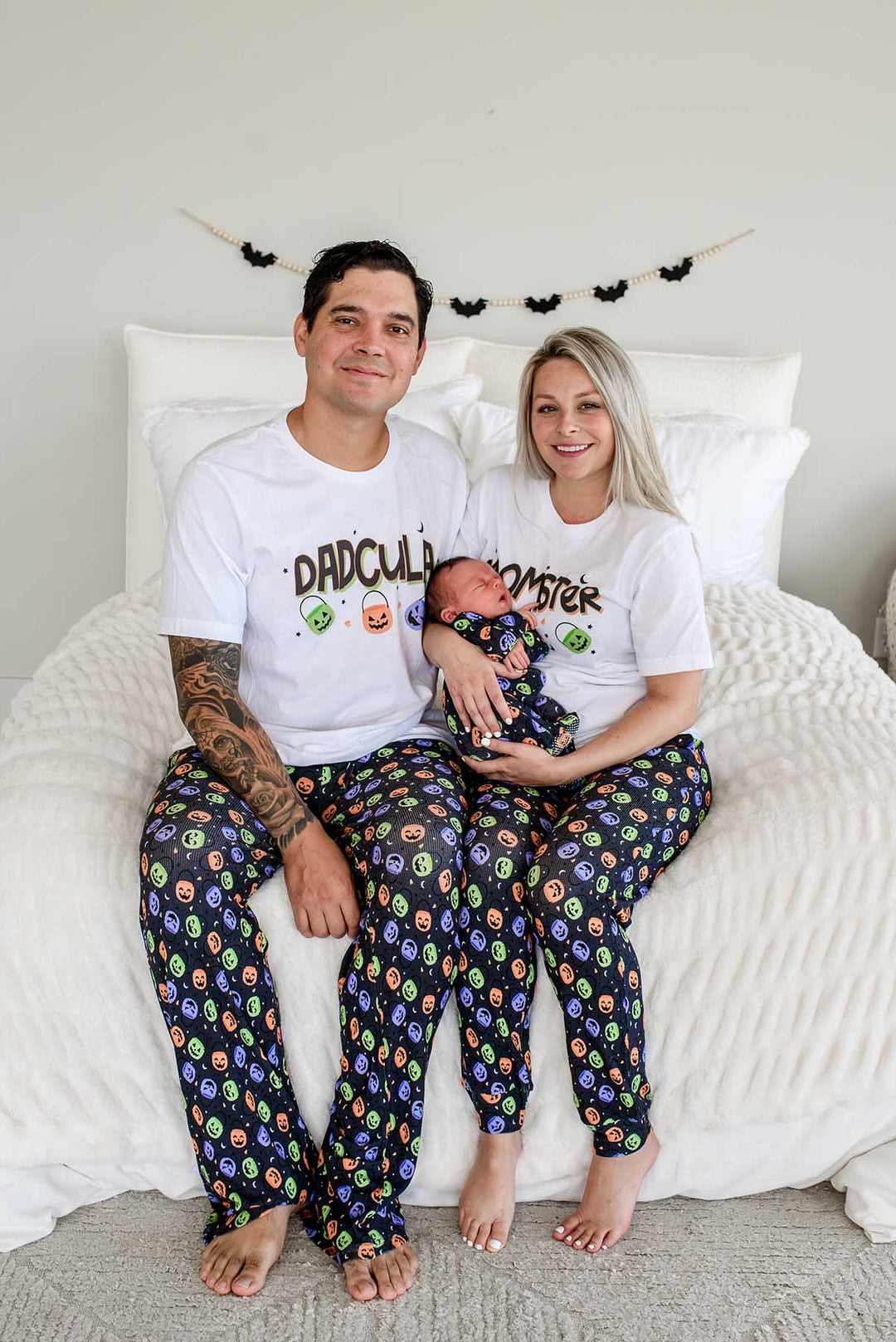 Father and mother sitting on bed wearing matching 'DADCULA' and 'MOMSTER' Halloween shirts with candy-print pajama pants, holding a newborn in matching print, bat garland on wall behind them