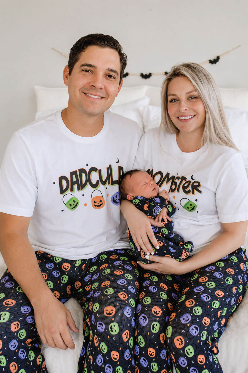 Family of three wearing matching Halloween Candy Crawl pajamas: father in 'DADCULA' t-shirt, mother in 'MOMSTER' t-shirt, both in candy-print pajama pants, holding a newborn swaddled in the matching print