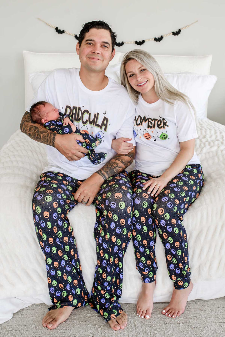 Family sitting on bed wearing matching Halloween pajamas: father in 'DADCULA' shirt holding newborn, mother in 'MOMSTER' shirt, both in candy-print pajama pants, bat garland on wall behind them