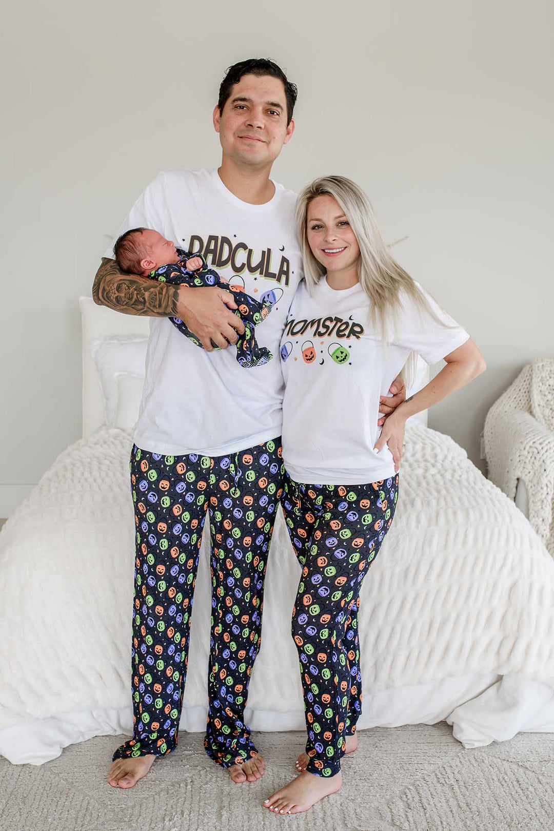 Father and mother standing beside bed wearing matching Halloween Candy Crawl pajamas: father in 'DADCULA' shirt holding newborn, mother in 'MOMSTER' shirt, both in candy-print pajama pants