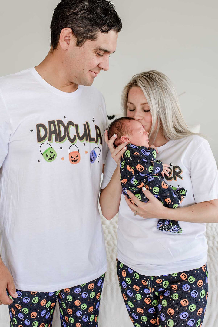 Father in 'DADCULA' t-shirt kissing newborn's head, mother in matching Halloween pajama top holding baby swaddled in candy-print blanket, both parents wearing candy-print pajama pants